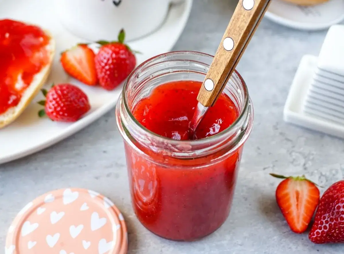 Ein Glas mit leuchtend roter Erdbeermarmelade, die mit einem Holzlöffel umgerührt wird. Daneben frische Erdbeeren und ein halbes Brötchen mit Marmelade – Erdbeermarmelade mit Pfiff für den perfekten Start in den Tag.