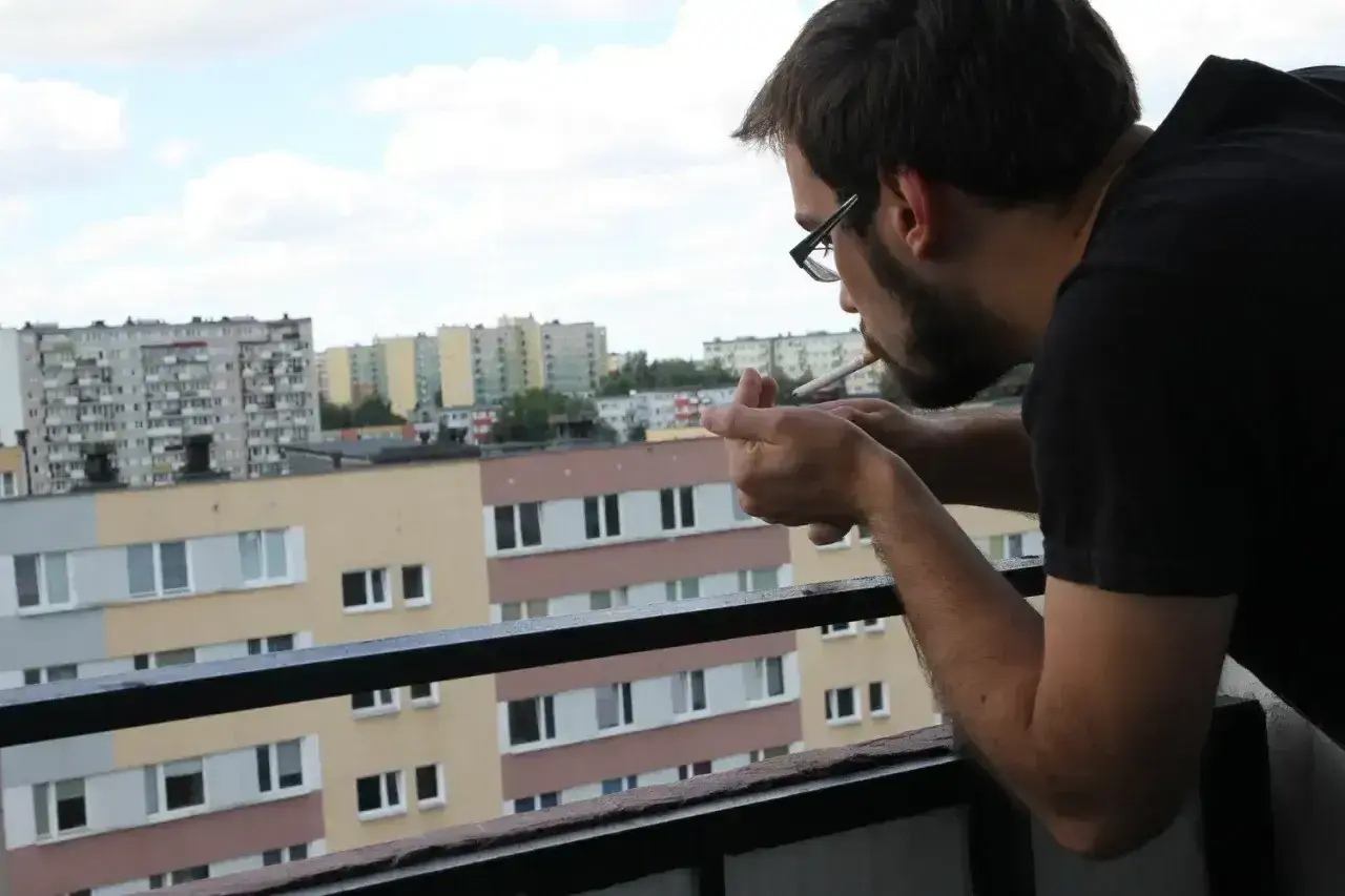 Mężczyzna zapala papierosa na balkonie, patrząc na blokowisko. Czy można palić na balkonie? To pytanie zadaje sobie wielu mieszkańców.
