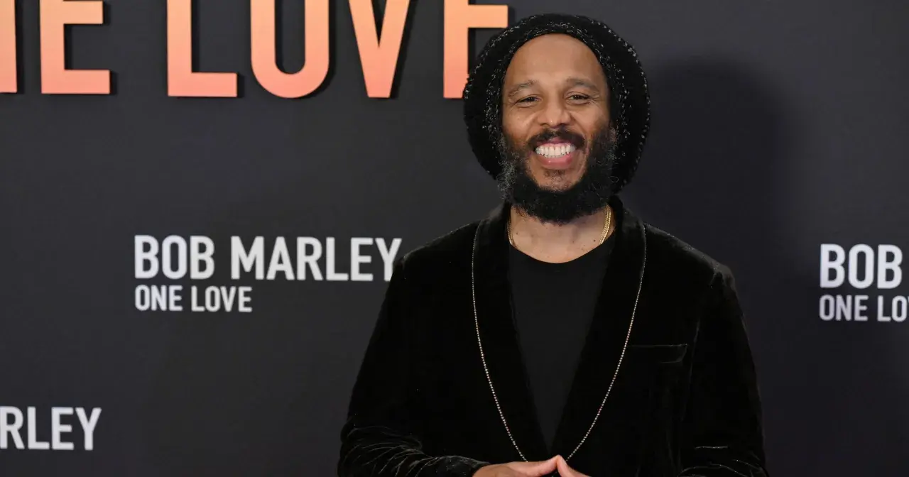 Uśmiechnięty mężczyzna w czapce i aksamitnej marynarce, przypominający Boba Marleya, z napisem "Bob Marley One Love" w tle. Może to być jeden z jego dzieci.