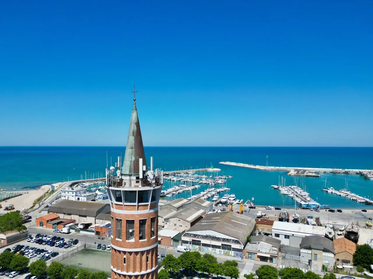 Torre con antenne domina il porto civitanova, pieno di barche e con il mare azzurro sullo sfondo.