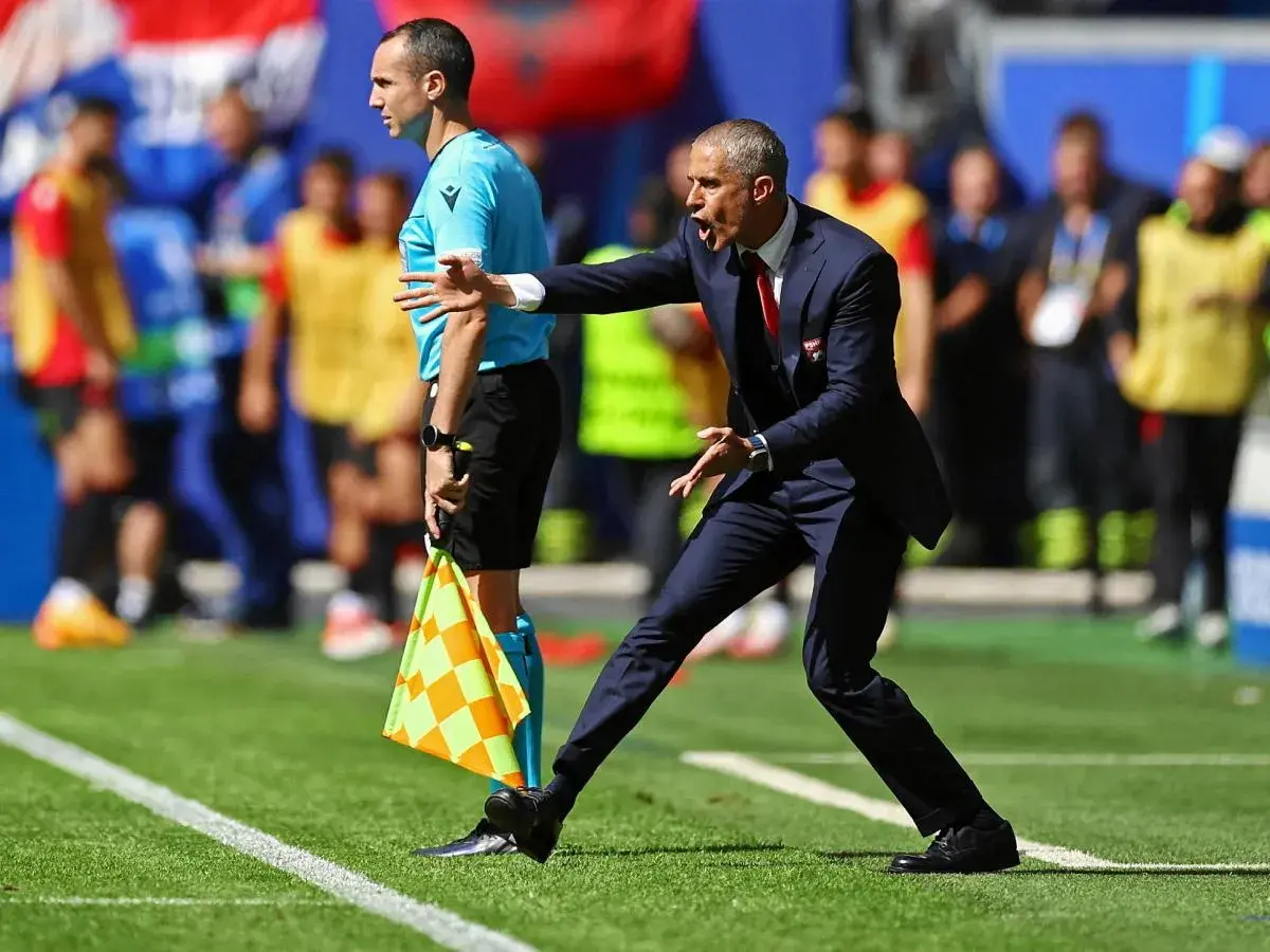 Sylvinho, trener w garniturze, gestykuluje na boisku. Obok stoi sędzia liniowy z flagą.
