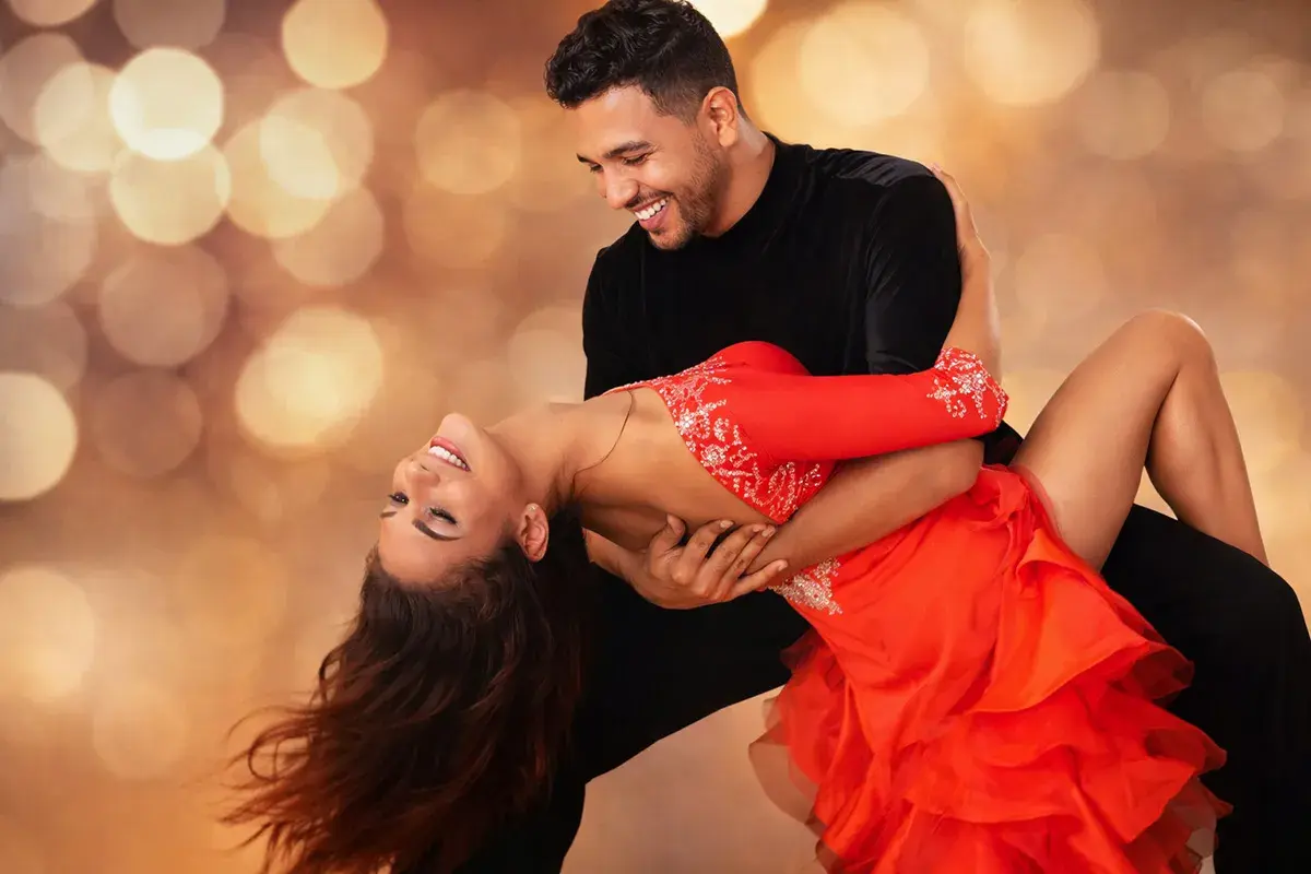 Coppia che balla con passione una salsa ballo latino, lei in abito rosso, lui in nero, su sfondo sfocato con luci dorate.