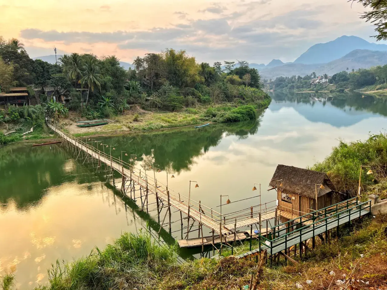 Mosty bambusowe nad rzeką, chatka z trawy, bujna zieleń i góry w oddali. To idealne miejsca, by zobaczyć **laos co zobaczyc**.