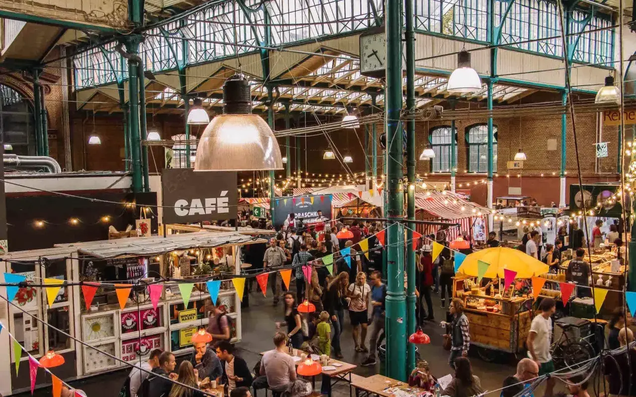 Buntes Treiben auf einem Street Food Festival in Berlin. Menschen schlendern zwischen Ständen mit Leckereien unter einer beeindruckenden Glasdachkonstruktion.