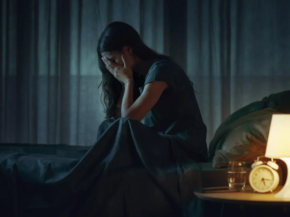 Mujer sentada en la cama, cubriéndose la cara con las manos, en plena vigilia del sueño. La noche es oscura, solo iluminada por una lámpara.
