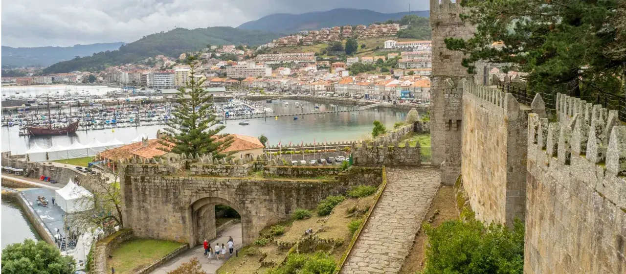 View from a castle wall overlooking a harbor filled with boats, a historic ship, and a town. The path leads through an ancient stone archway, hinting at a journey from porto to Baiona.
