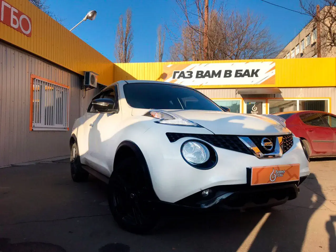 Biały Nissan Juke 1.6 benzyna z czarnymi felgami przed budynkiem z żółtym banerem "GAZ WAM W BAK".