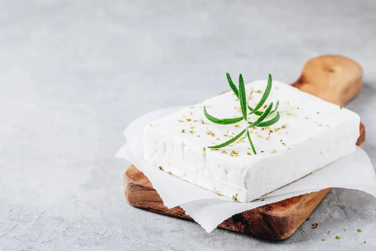 Un blocco di feta su un tagliere di legno, guarnito con rosmarino e spezie. Ricorda che la feta contiene lattosio.