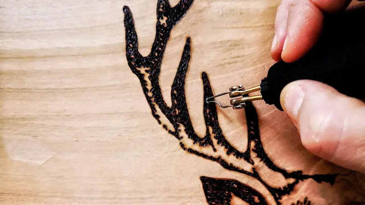 Dettaglio di lavori con pirografo su legno: una mano che disegna un palco di cervo con precisione.