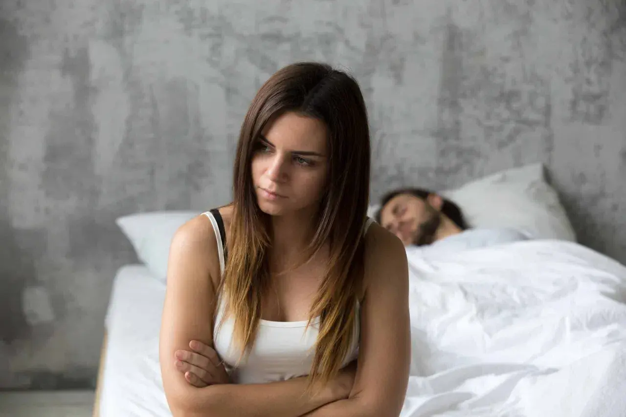 Een vrouw zit met gekruiste armen, gefrustreerd, terwijl haar partner slaapt. Ze probeert zich in te leven in anderen, maar voelt zich alleen.