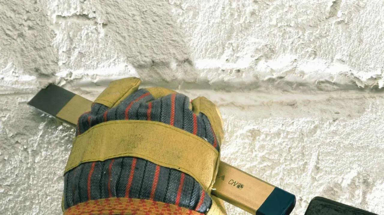 Mit einem Meißel wird alter **Putz entfernt**. Eine Hand mit Arbeitshandschuh hält das Werkzeug.