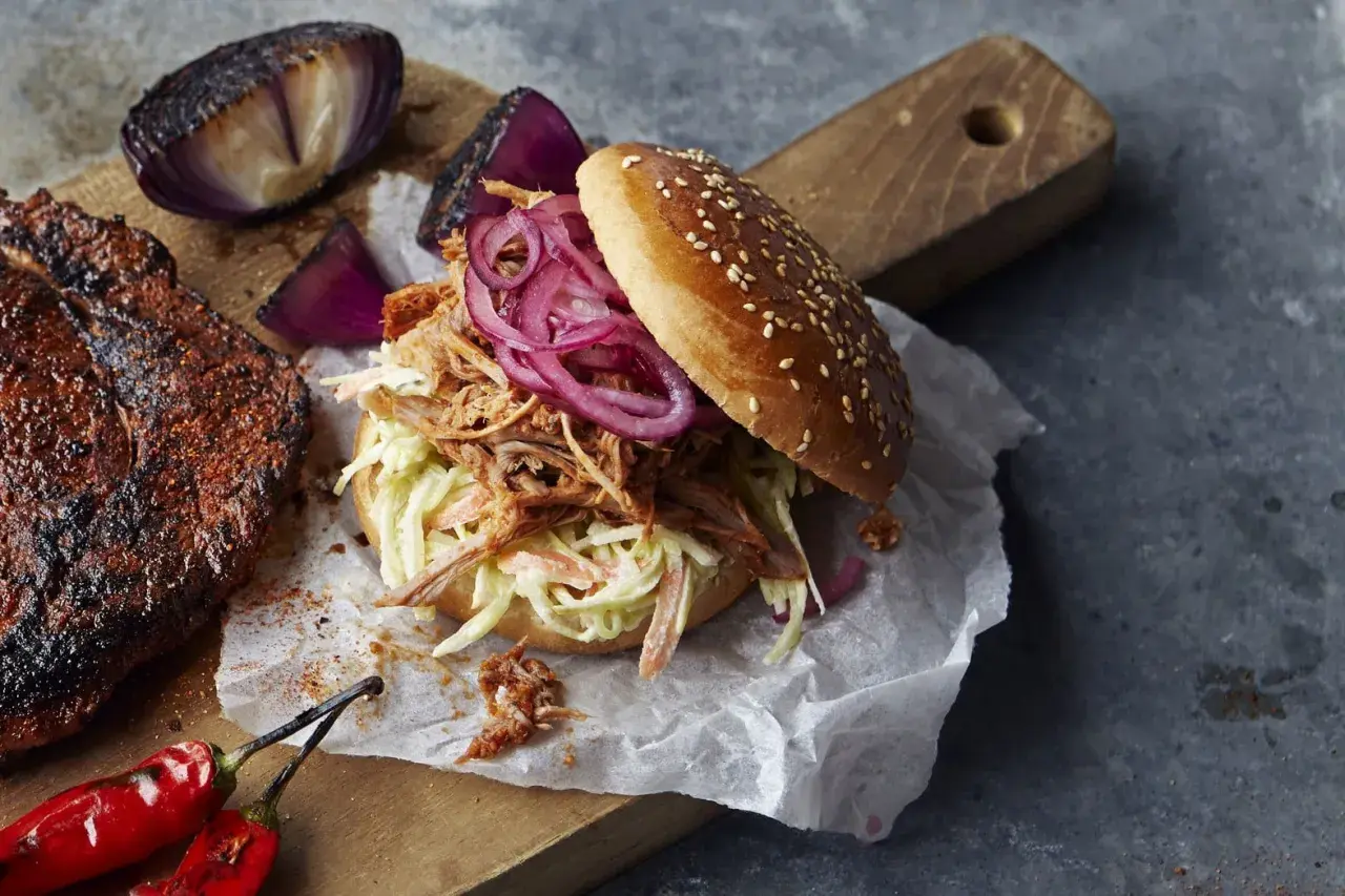En saftig pulled pork-burgare med krispig coleslaw och rödlök, perfekt för ett bbq-recept. Bredvid ligger en bit grillat kött och rostade lökar.