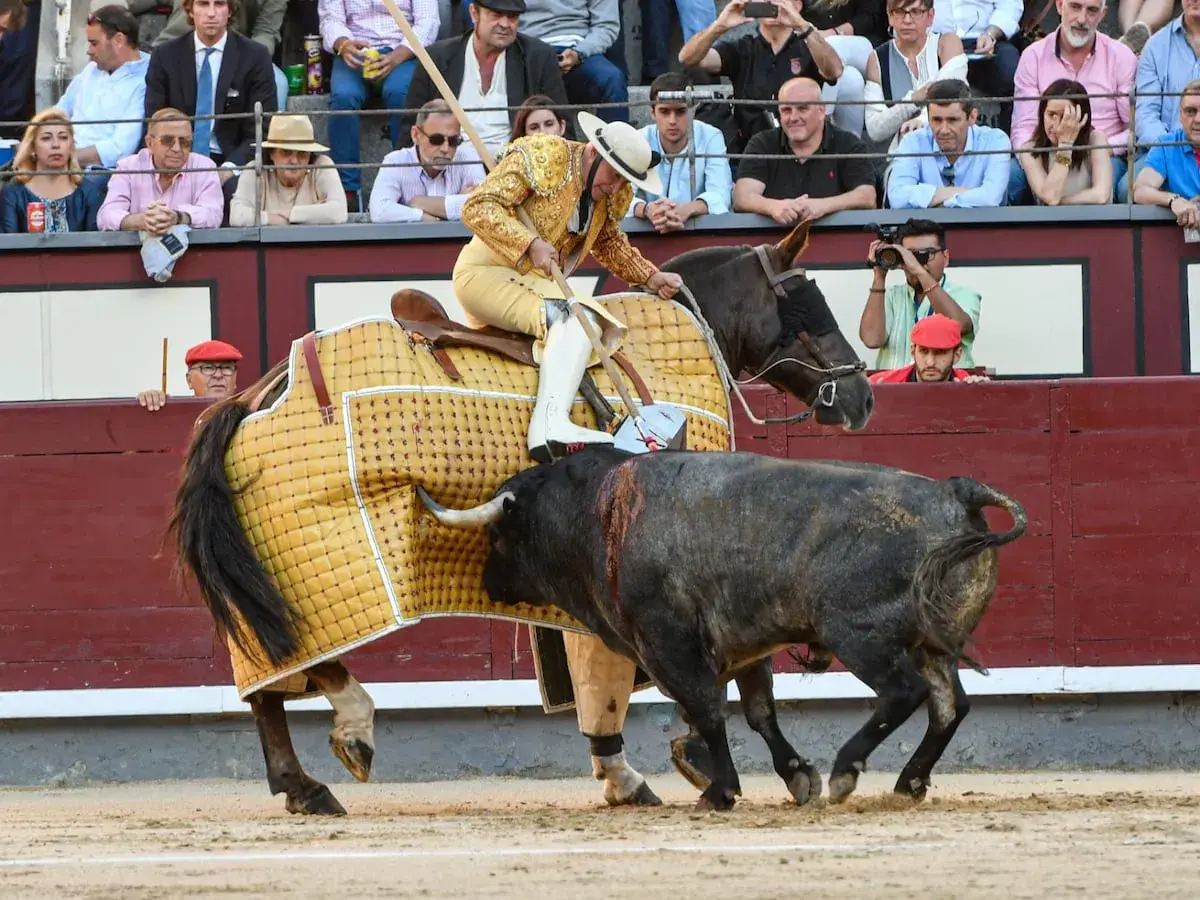 Un torero a caballo, con traje de luces, se enfrenta a un toro en la arena. El caballo lleva una manta amarilla. El público observa desde las gradas.
