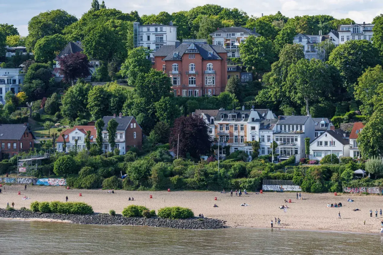Prächtige Villen und grüne Hügel über einem Sandstrand an den Elbvororten Hamburgs. Menschen genießen den sonnigen Tag am Wasser.