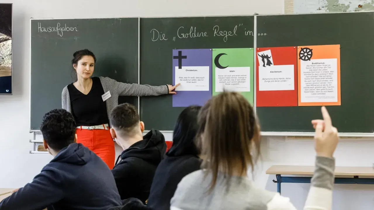Lehrerin erklärt die Goldene Regel im Religionsunterricht Grundschule. Tafeln zeigen Zitate aus Christentum, Islam, Alevitentum und Buddhismus.