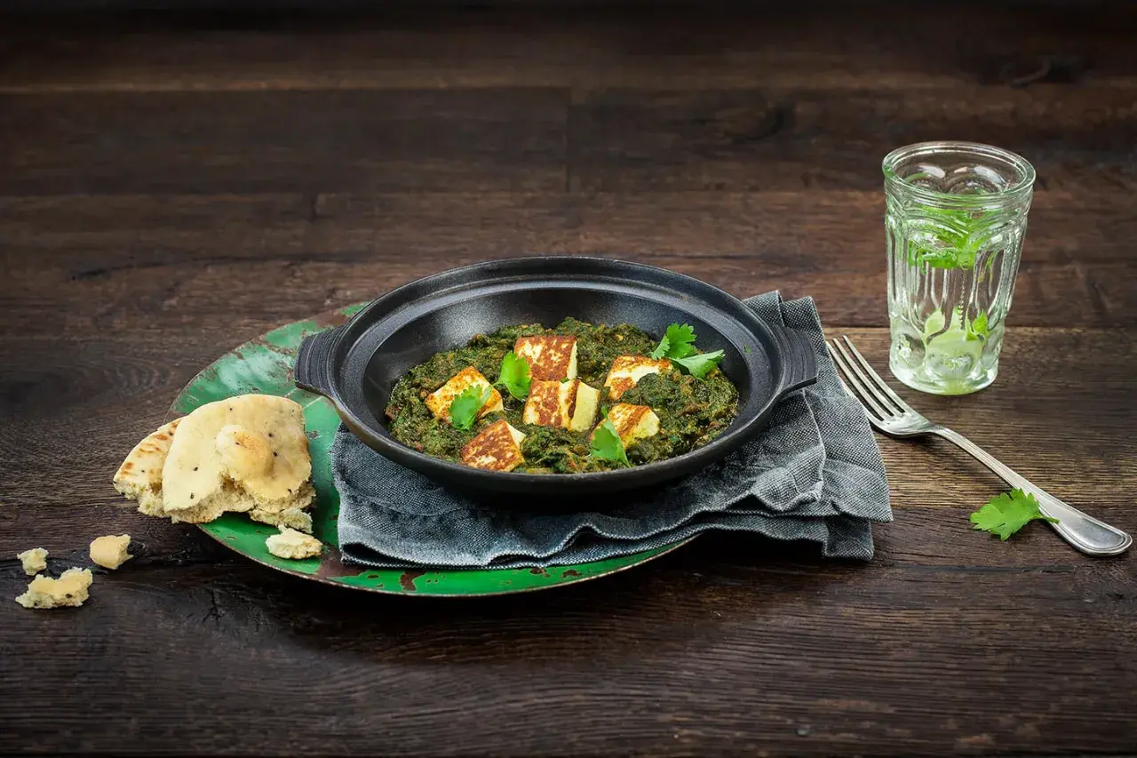 En skål med krämig palak paneer, toppad med gyllene paneerbitar och färsk koriander. Serveras med naanbröd och en glasdryck.