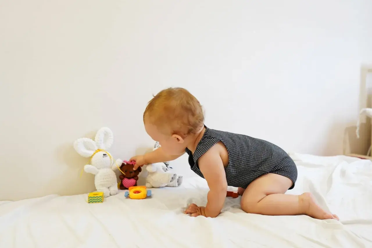 A baby explores Montessori baby activities, reaching for a teddy bear among soft toys on a white blanket.
