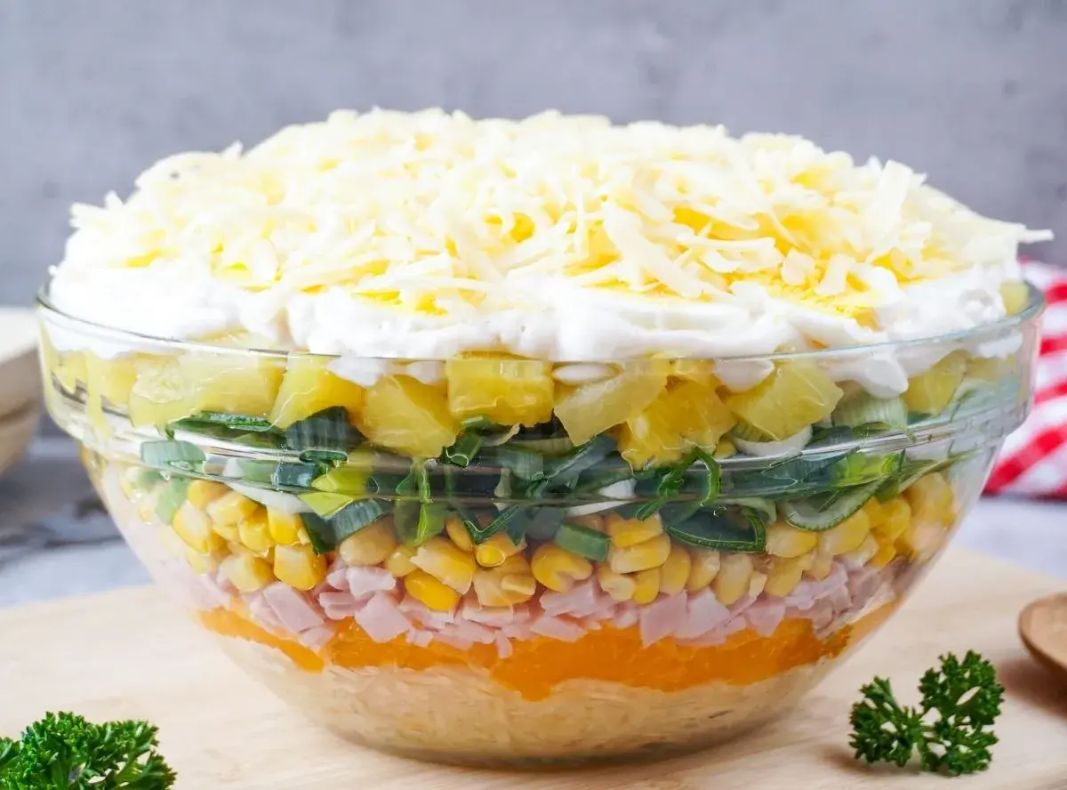 Schichtsalat mit Eisbergsalat, Mais, Schinken, Ananas, Lauch und Käse in einer Glasschüssel.
