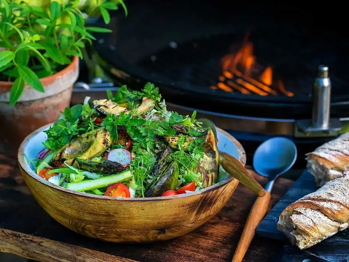 En stor skål med god sallad, grillad aubergine och sparris, med en grill i bakgrunden. Perfekt för en sommarkväll.