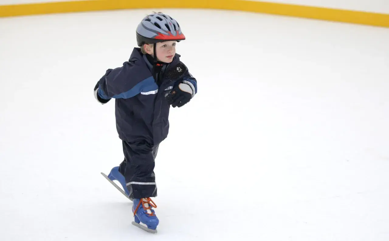 Bambino con casco e tuta blu pratica il pattinaggio sul ghiaccio, con i pattini azzurri e lacci arancioni.
