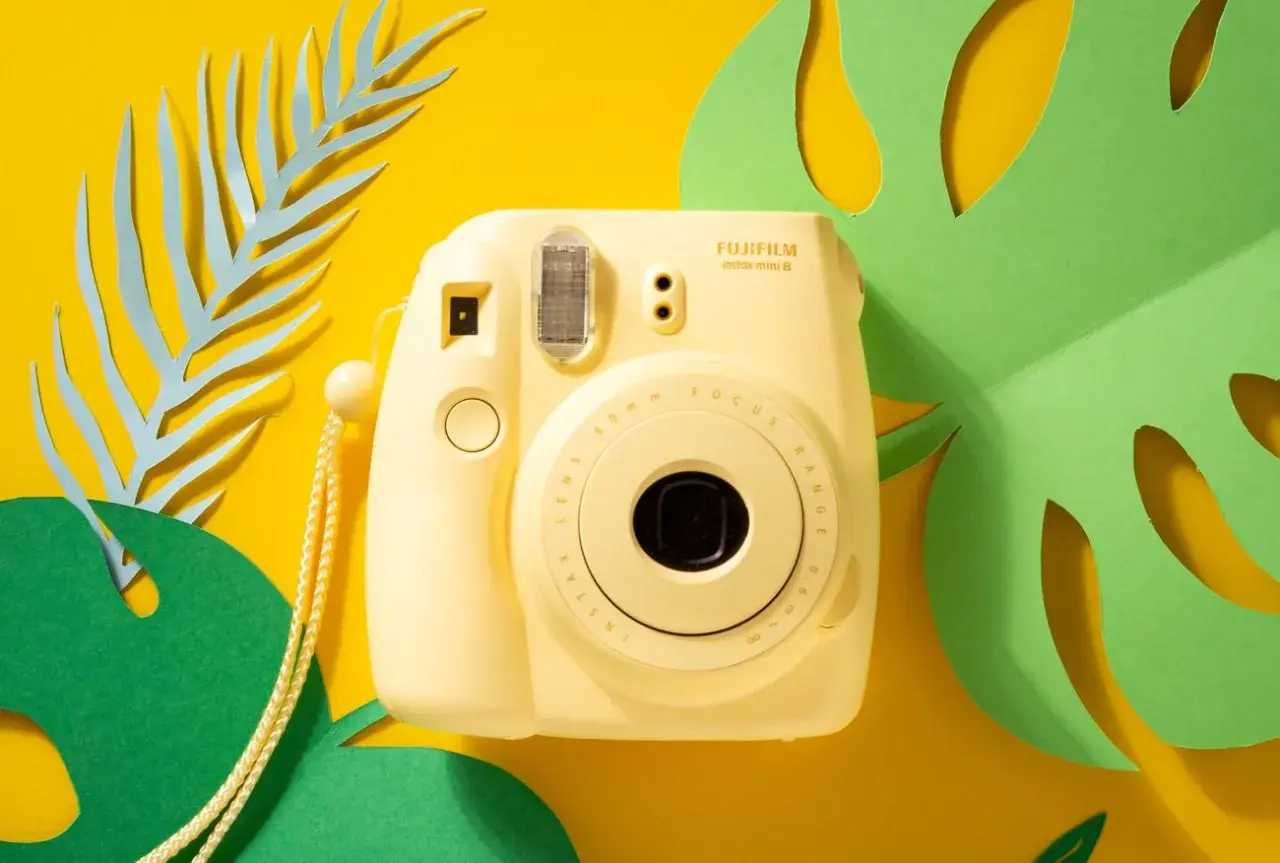 Żółty aparat natychmiastowy Fujifilm Instax Mini 8 na tle żółtym z zielonymi i niebieskimi liśćmi. Idealny do wakacyjnych zdjęć, często pojawia się w rankingach aparatów natychmiastowych.