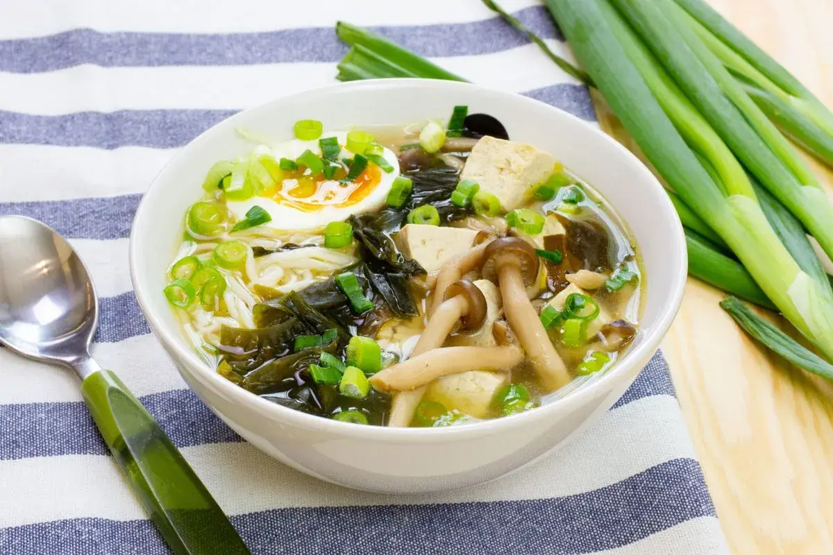 A miska zupy miso z tofu, jajkiem na pół, grzybami i szczypiorkiem. Obok leży zielona łyżka i pęczek dymki.