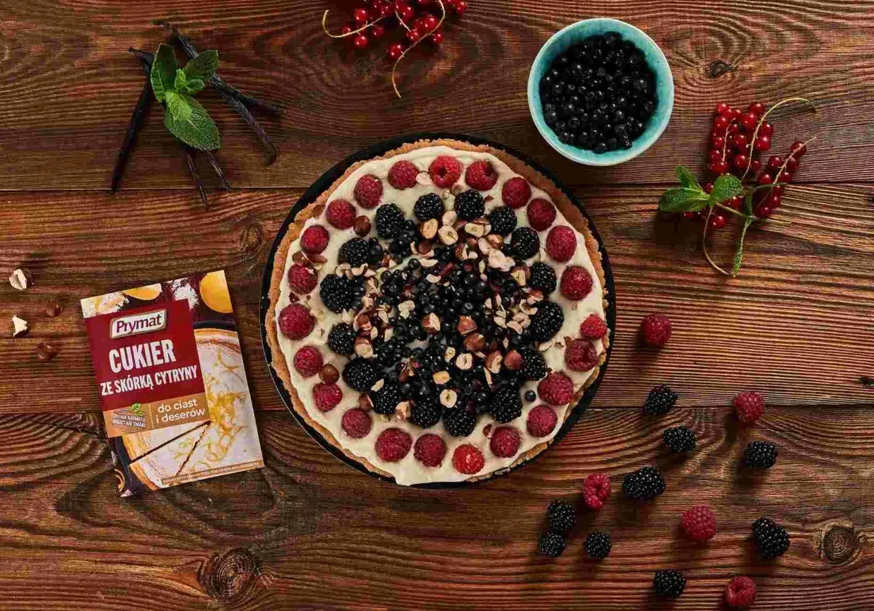 Pyszna tarta z mascarpone, malinami, jeżynami i borówkami, posypana orzechami. Obok opakowanie cukru z skórką cytrynową.