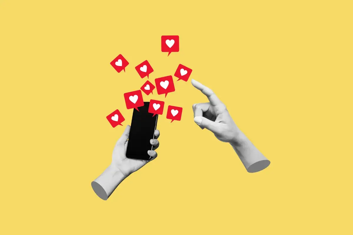 Dwie ręce, jedna trzyma smartfon, druga wskazuje na ekran, z którego wylatują czerwone dymki z serduszkami, symbolizujące polubienia w social mediach.