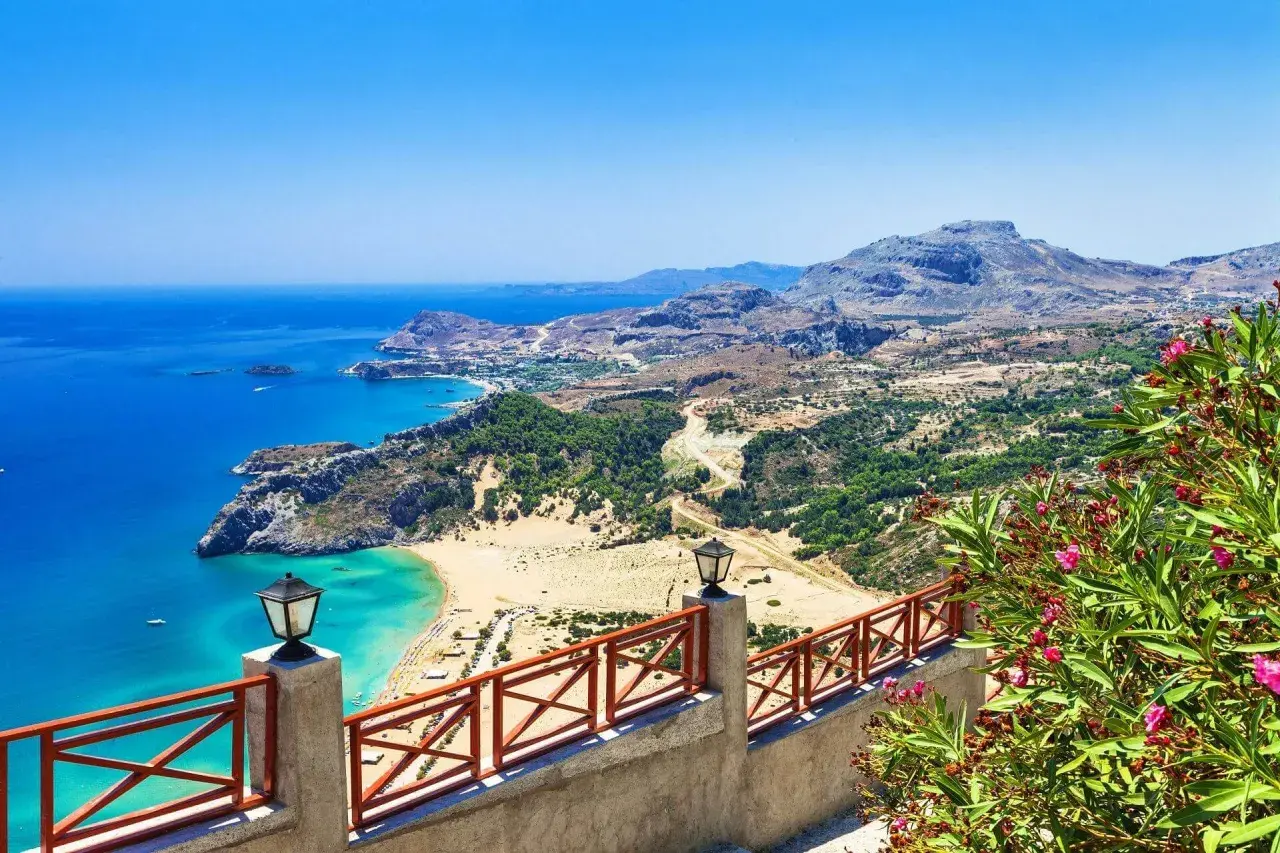 Blick auf eine Bucht mit türkisfarbenem Wasser, Sandstrand und grünen Hügeln. Ein Geländer mit Laternen und blühenden Oleandern im Vordergrund. Rhodos Geheimtipps für unvergessliche Ausblicke.