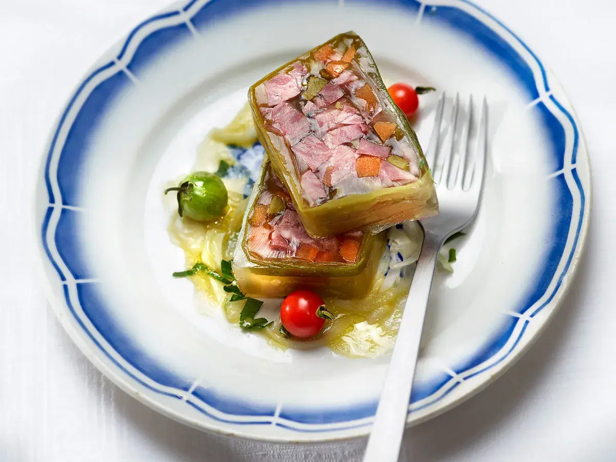 Deux portions de fromage de tête translucide, garnies de légumes et de viande, sur une assiette ornée de bleu.