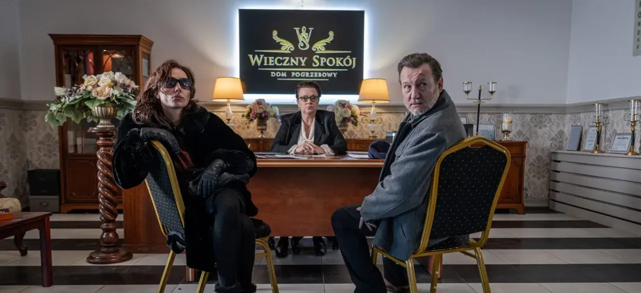 Trzy osoby siedzą w eleganckim biurze domu pogrzebowego. W tle logo "Wieczny Spokój". Scena z serialu "Minuta ciszy sezon 2".