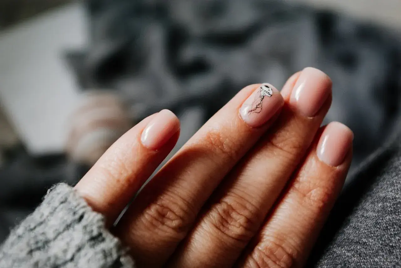 Une main aux ongles rose pâle, avec une délicate méduse dessinée sur l'annulaire. Une manucure pastel subtile et élégante.