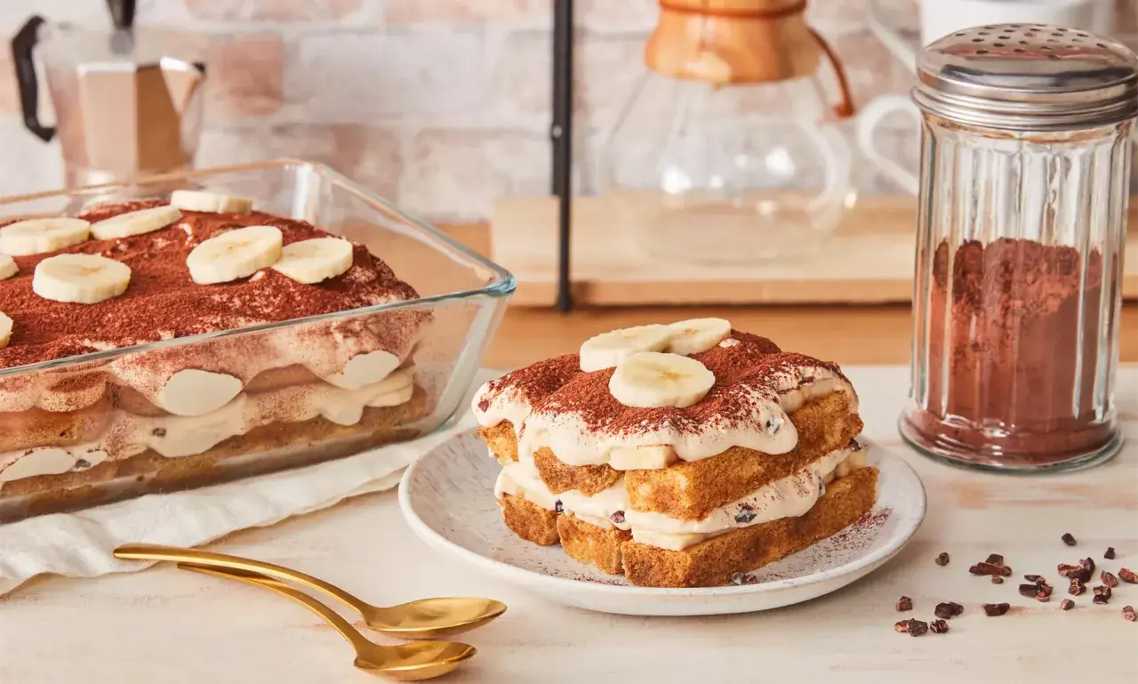 Ein köstliches Bananen Tiramisu, geschichtet mit Creme und Bananenscheiben, bestäubt mit Kakao. Daneben ein Glas Kakaopulver.