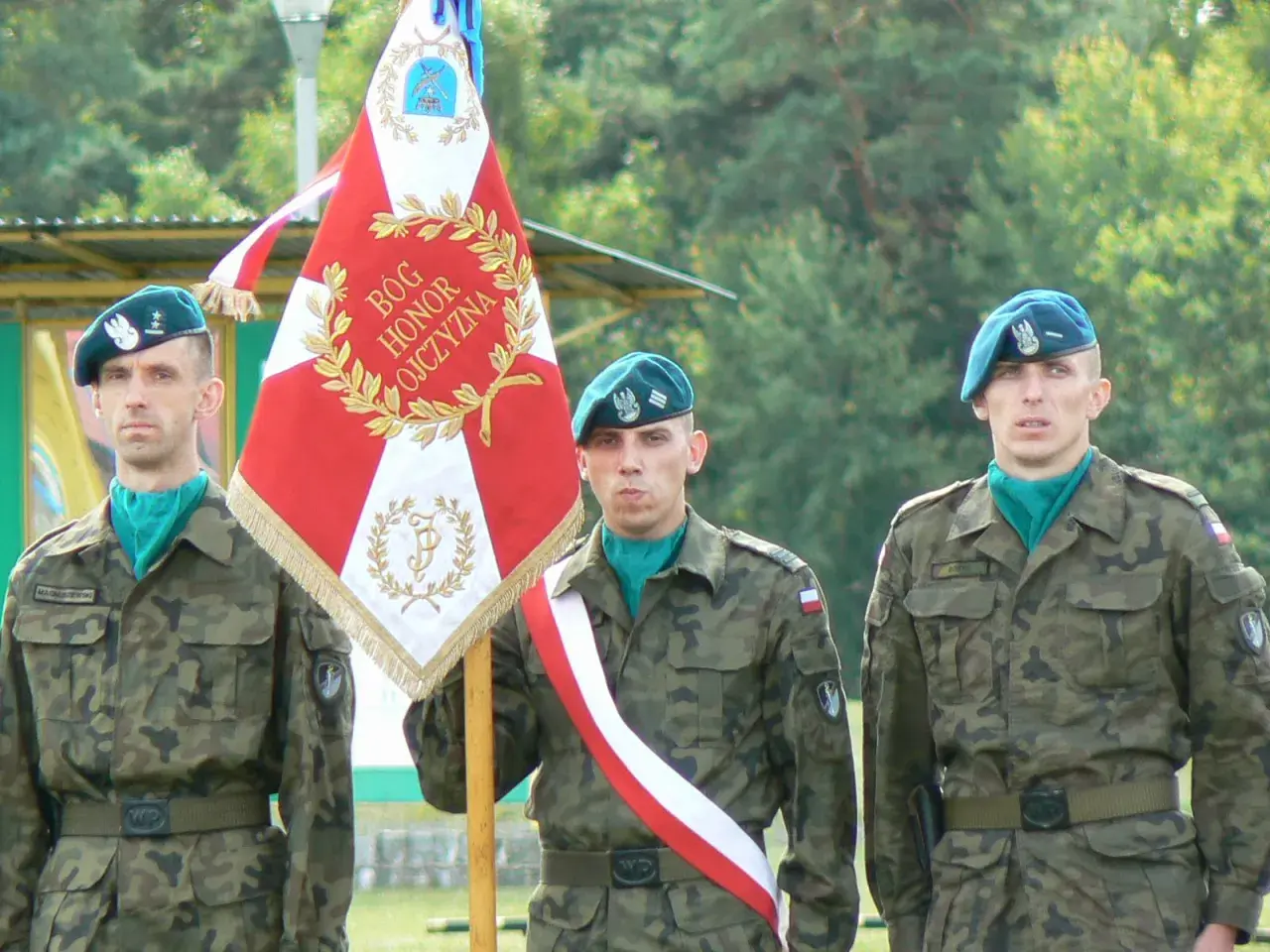 Trzech żołnierzy rezerwy w mundurach i beretach stoi obok sztandaru.