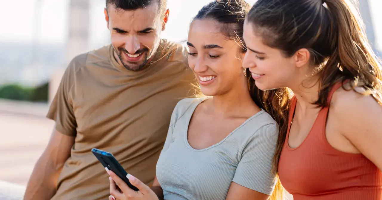 Trzech przyjaciół ogląda coś na telefonie. Uśmiechnięta dziewczyna, która trzyma telefon, może ukrywać zakochanie, gdy patrzy na ekran.