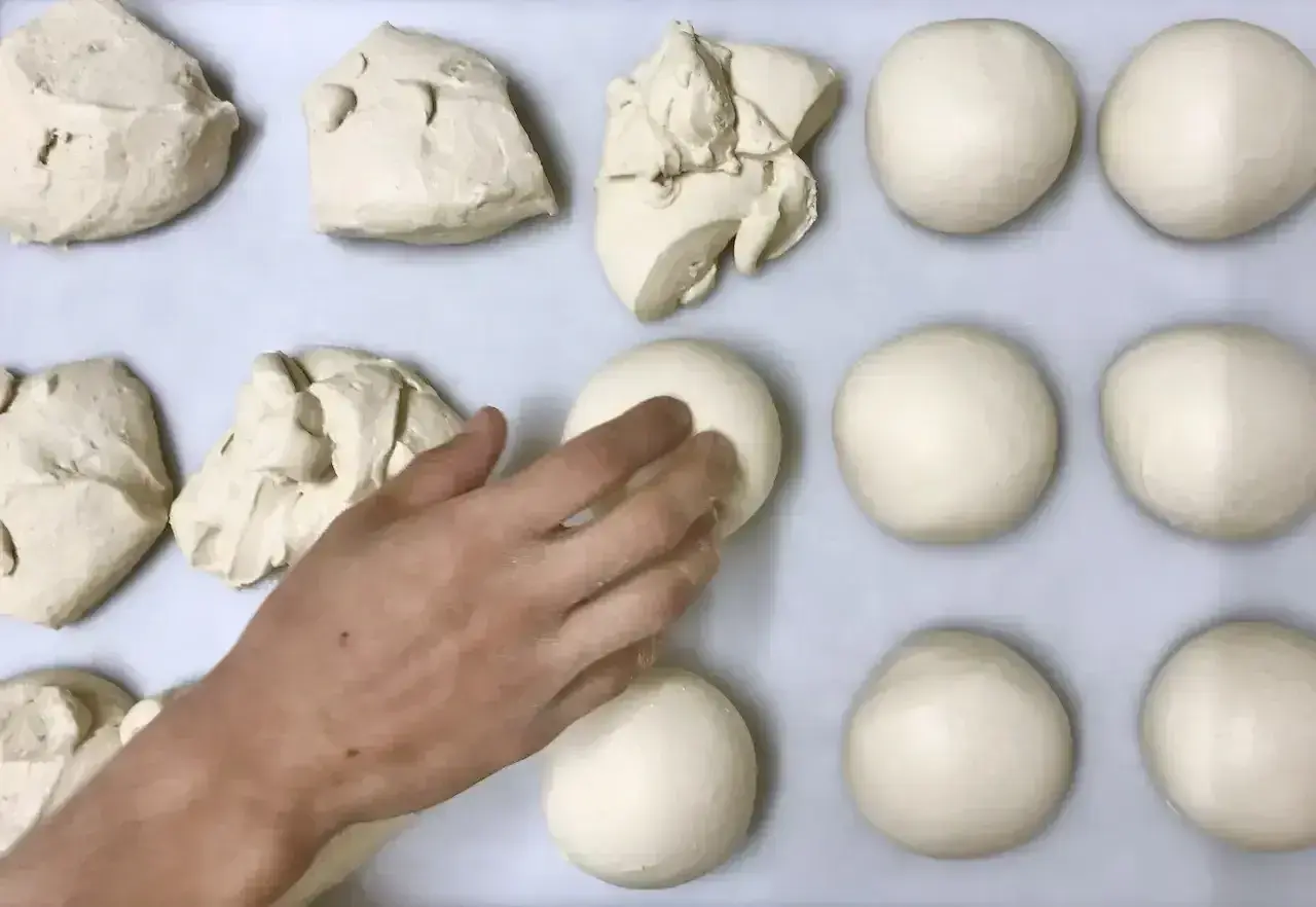 Des boules de pâte à pizza sont disposées sur une plaque. Une main façonne délicatement l'une d'elles.