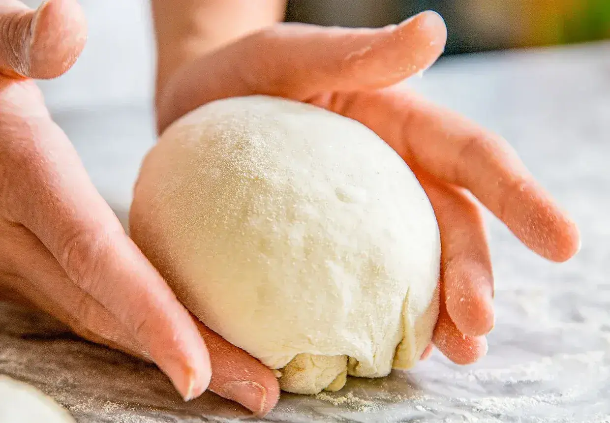 Hände formen einen Teigball auf einer bemehlten Oberfläche.