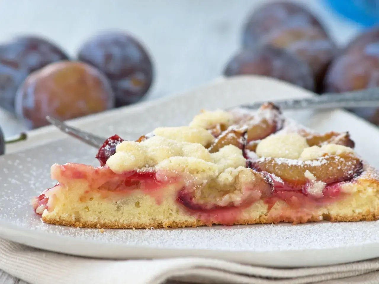Ein saftiges Stück Apfel Pflaumen Kuchen mit Streuseln, bestäubt mit Puderzucker. Im Hintergrund sind frische Pflaumen zu sehen.