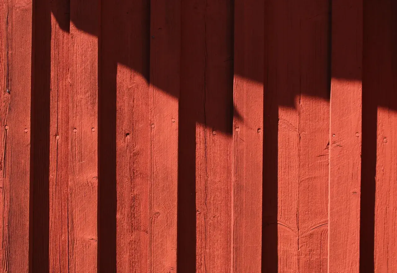 Röd träfasad med skuggor som faller som vertikala streck. Träet är målat tunt, vilket ger en rustik känsla.