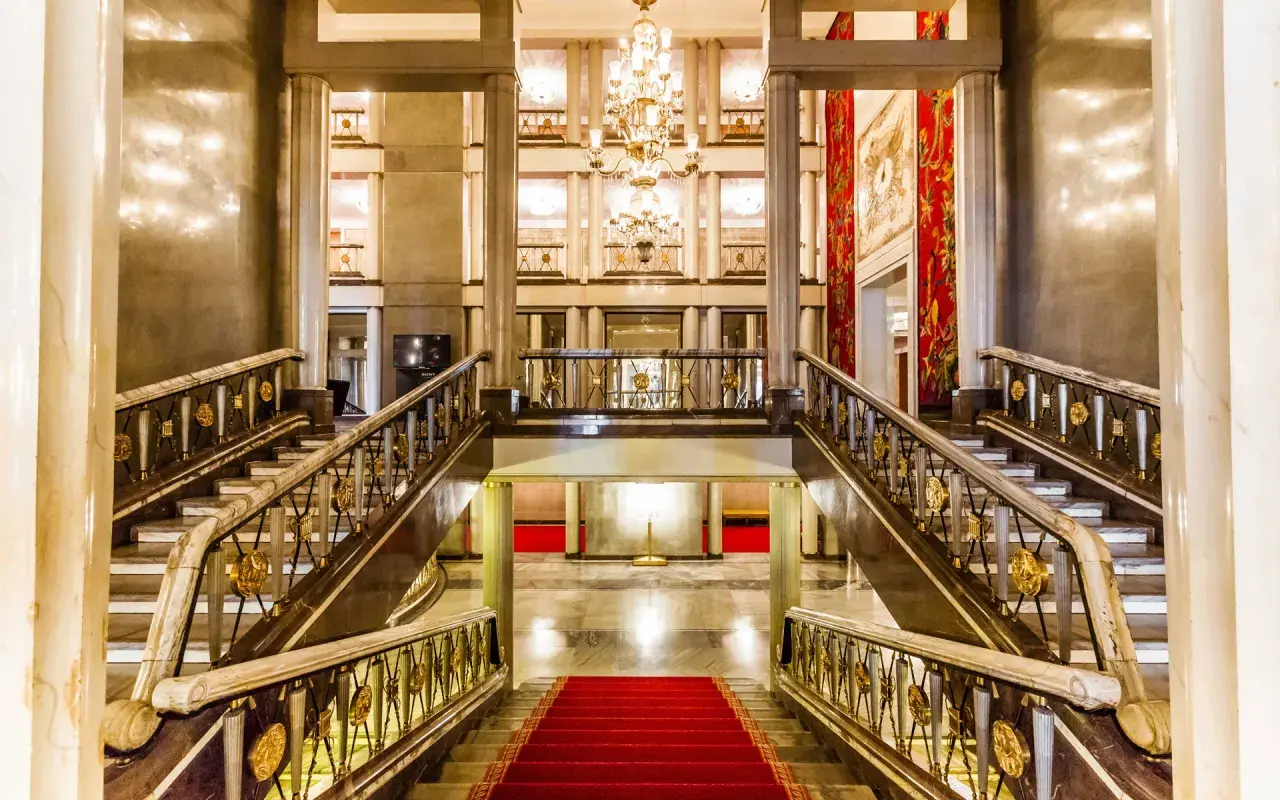 Elegancki foyer z marmurowymi schodami, złotymi balustradami i kryształowym żyrandolem. Czerwony dywan prowadzi do przestronnego holu.