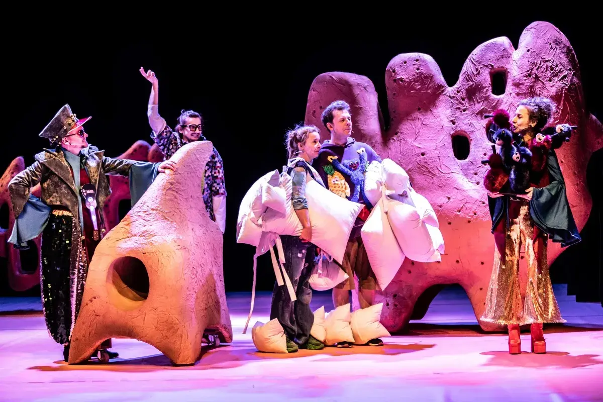 Aktorzy w barwnych strojach, niosący poduszki, na tle scenografii przypominającej gigantyczne, różowe formacje. To fragment spektaklu z repertuaru teatru lalki.