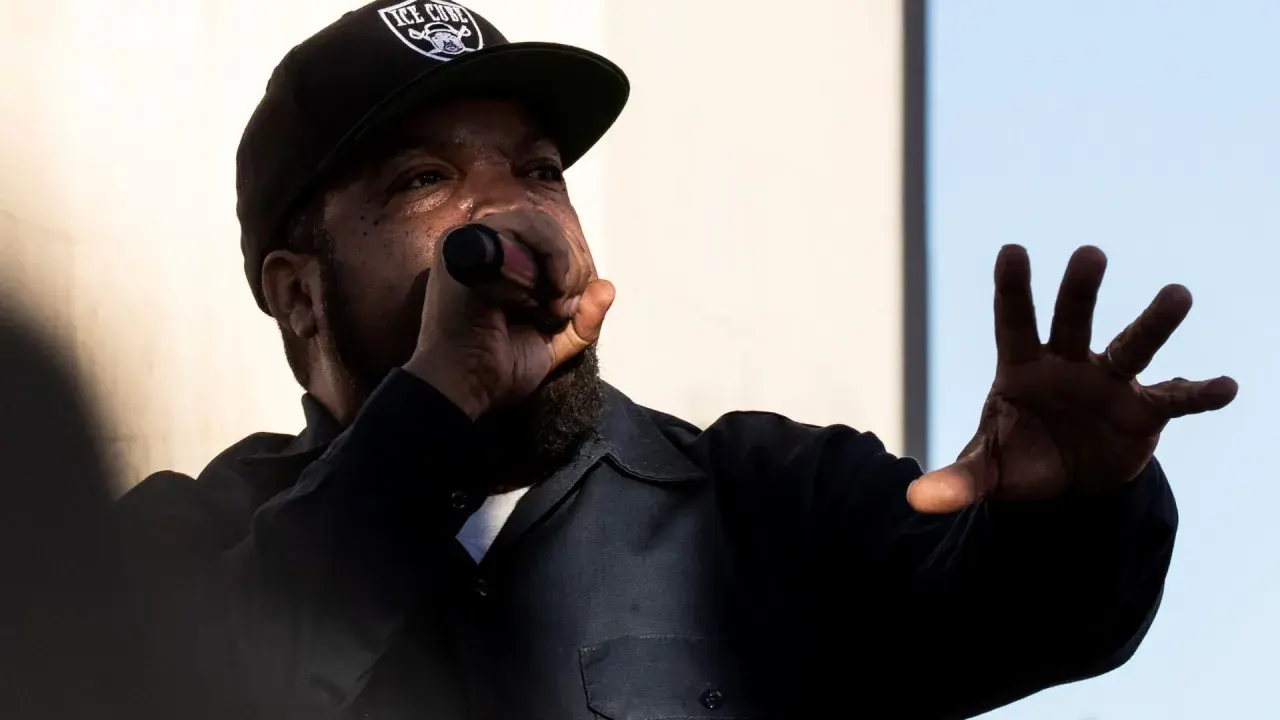Raper Ice Cube w czapce z daszkiem "ICE CUBE" śpiewa do mikrofonu, gestykulując ręką. Jego styl to klasyczny diss.