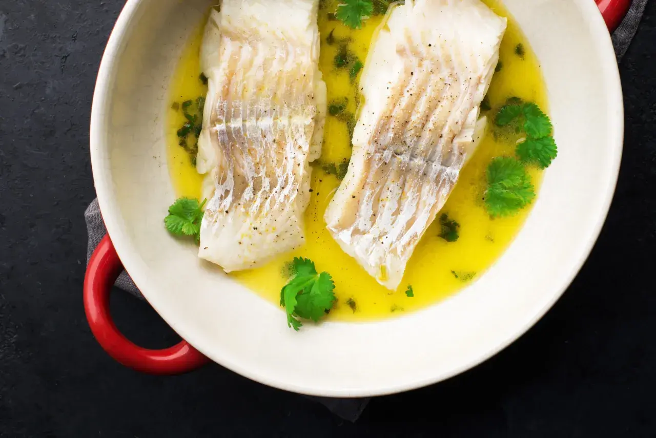 Dwa filety halibut pieczony ze skórą w maśle z ziołami i kolendrą, podane w białej misie z czerwonymi uchwytami.