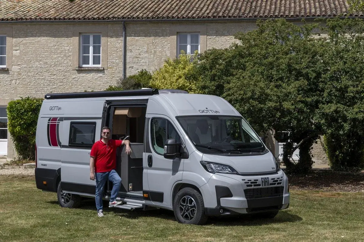 Un homme pose devant un **fourgon ou camping car** gris, porte ouverte, devant une maison en pierre et des arbres.