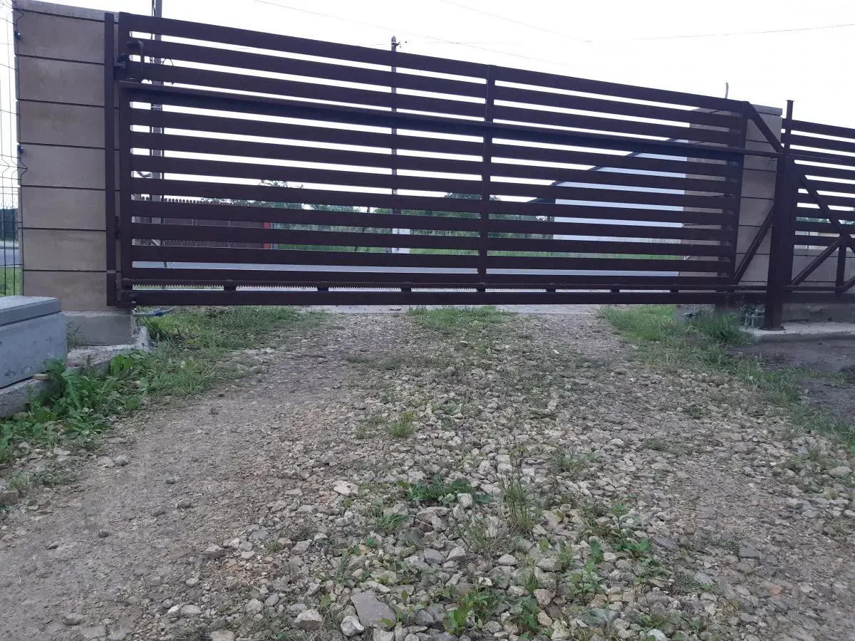 Brązowa, metalowa brama przesuwna z poziomymi szczeblinami. Widoczna jest szczelina pod bramą, przez którą widać trawę i żwir.