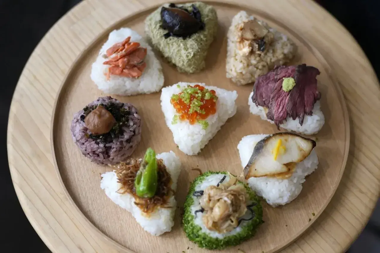 A colorful assortment of Japanese triangle food, including onigiri with salmon, beef, fish, and ikura, arranged on a wooden platter.