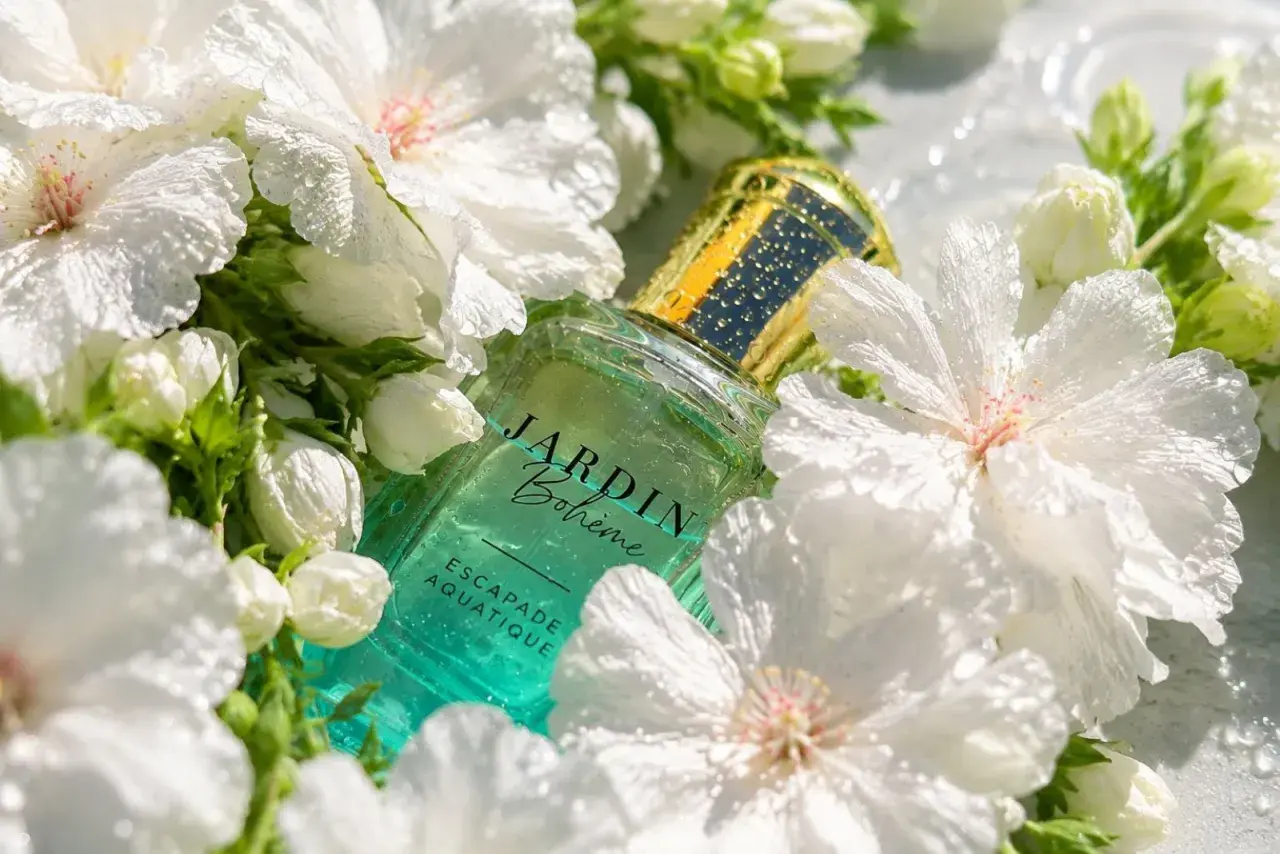 Szklana butelka perfum Jardin Bohème Escapade Aquatique, otoczona białymi kwiatami. Ten zapach to jak szczęśliwy los na lato.
