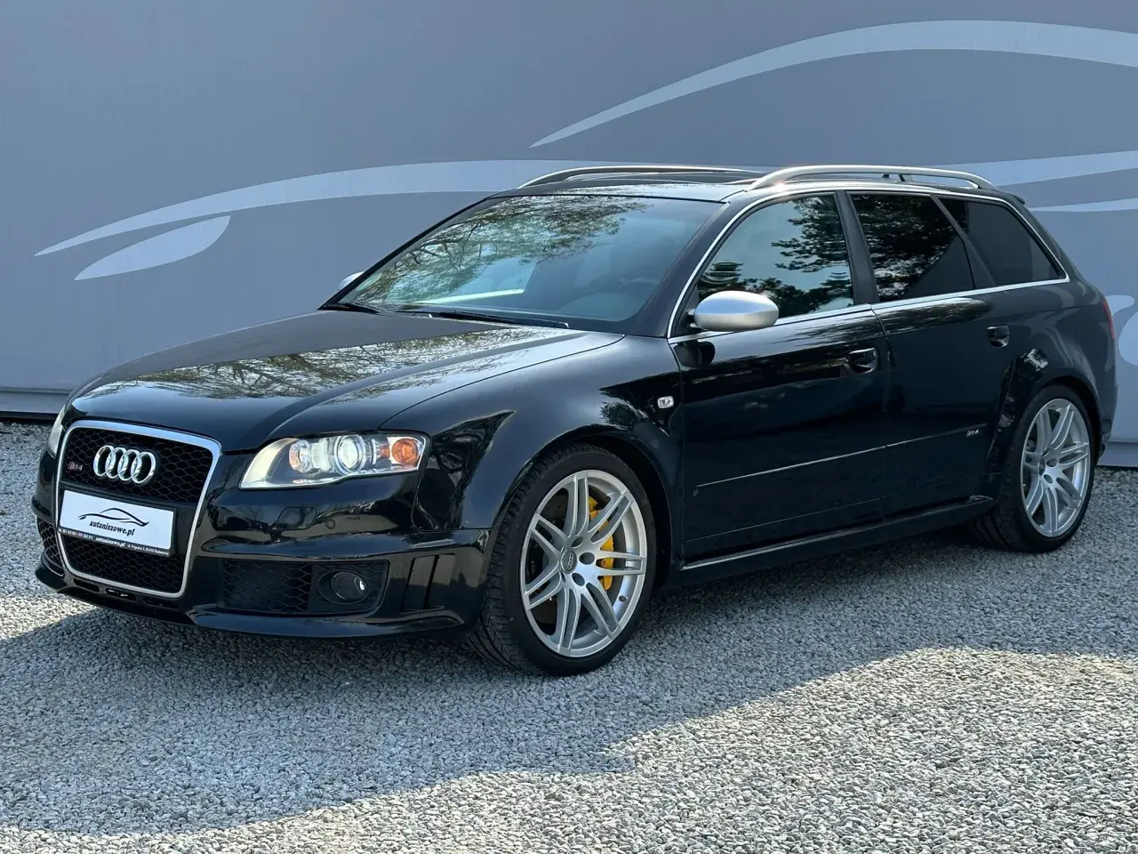 Czarny Audi RS4 B7 Avant z żółtymi zaciskami hamulcowymi, zaparkowany na żwirze przed szarą ścianą z grafiką.