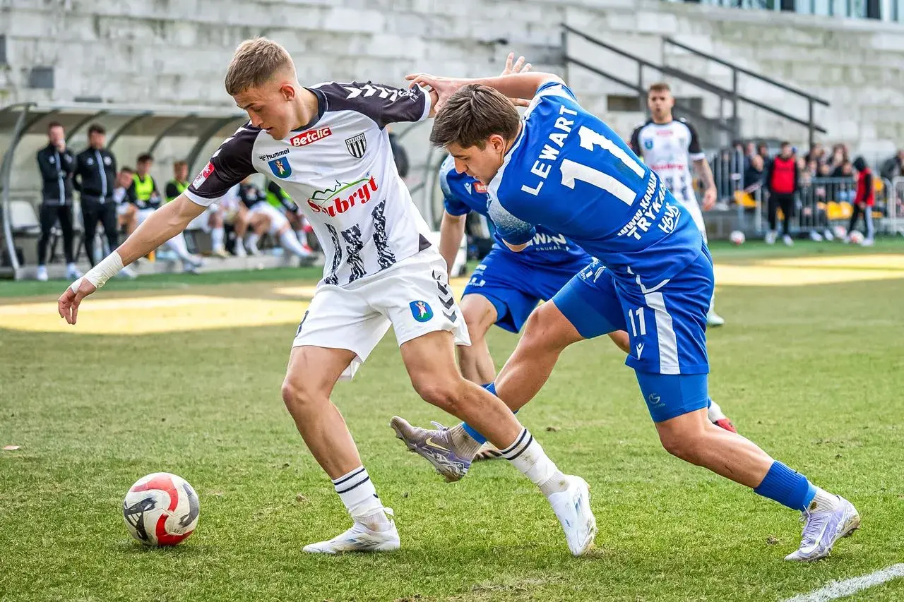 Piłkarze walczą o piłkę na boisku. Jeden w białej koszulce, drugi w niebieskiej z numerem 11. To ważny mecz, który może wpłynąć na 3. liga tabelę.