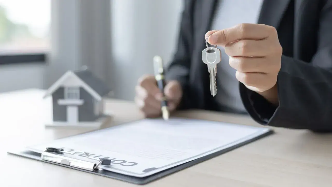 Persona firma un contrato de alquiler con opción a compra, sosteniendo la llave de una casa modelo.